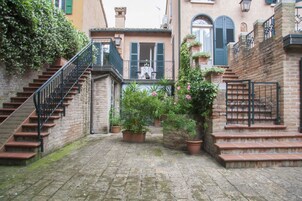Un patio estrecho con una escalera de piedra que lleva a un balcón en el segundo piso, rodeado de macetas con plantas y un muro de ladrillo.