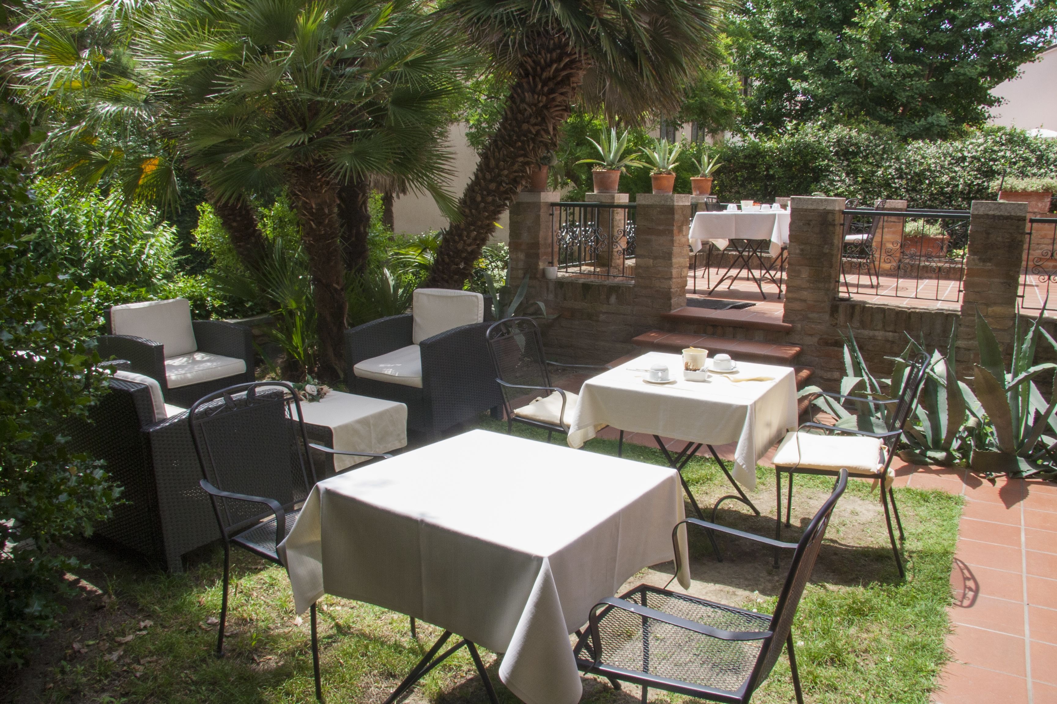 Patio esterno con tavoli e sedie, immerso nel verde e circondato da palme.