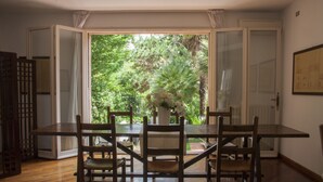 Un comedor con mesa y sillas de madera, un jarrón con flores y vista a zonas verdes a través de puertas abiertas.