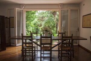 Un comedor con mesa y sillas de madera, un jarrón con flores y vista a zonas verdes a través de puertas abiertas.