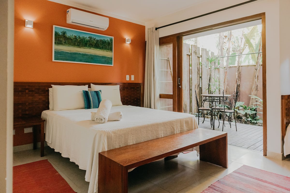 A bedroom with a large bed, a wooden bench, a painting of a beach, and a view of a patio with plants and a table.