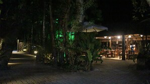 Un'area esterna illuminata di notte, con un vialetto lastricato, una zona coperta con tavoli e sedie e vegetazione rigogliosa.