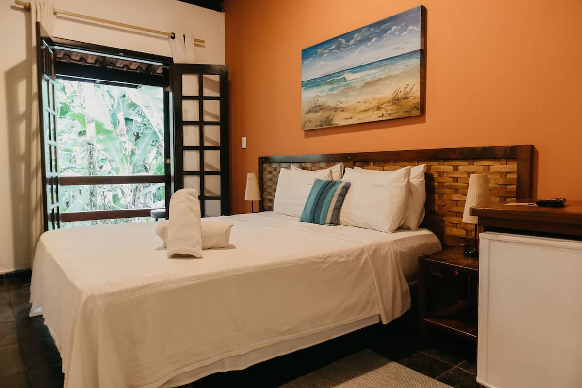 A neatly made bed with white linens and a wooden headboard, a painting of a beach scene on the wall, and a window with a view of greenery.