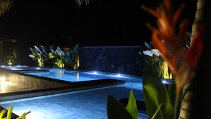 Area piscina illuminata di notte, con bordi luminosi, circondata da vegetazione rigogliosa e un solarium piastrellato.