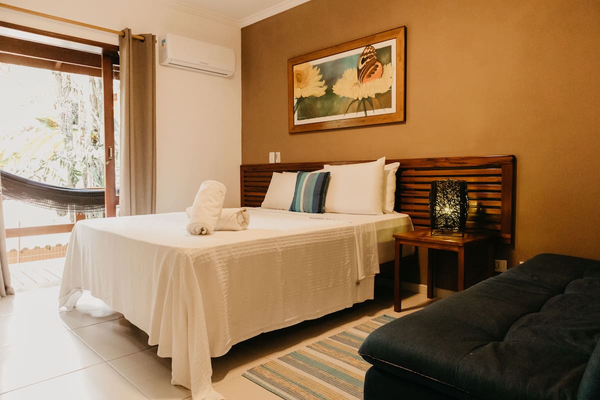 A bedroom with a bed, a nightstand, a lamp, a sofa, a hammock, and a painting on the wall.