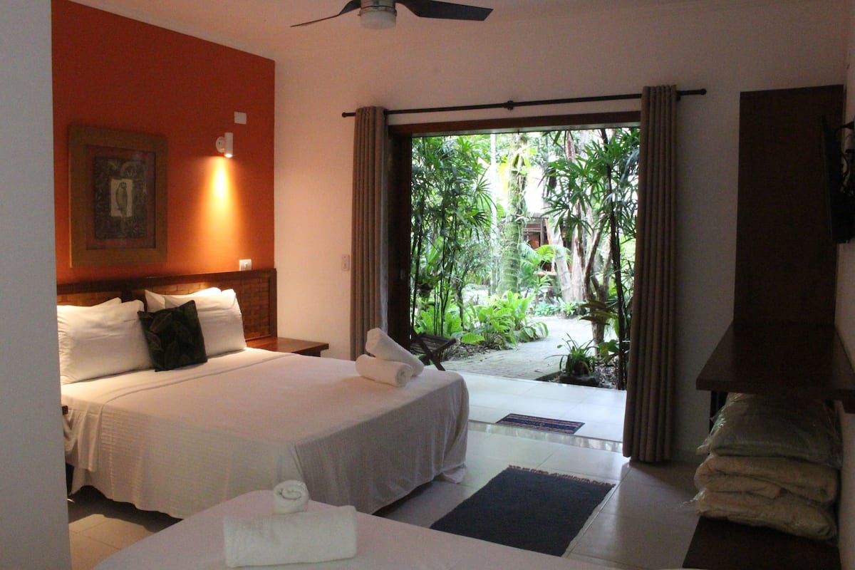 A hotel room with a bed, a ceiling fan, a view of greenery outside, and a door leading to a balcony.