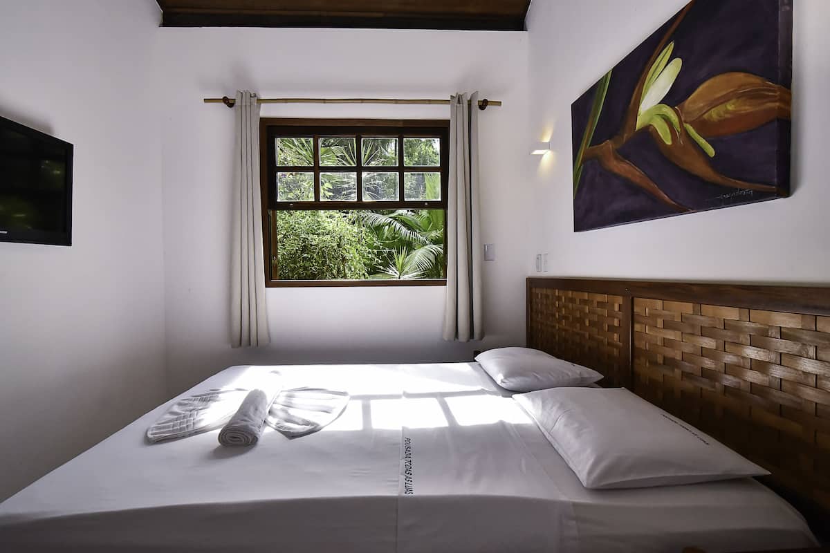 A neatly made bed with white linens and a rolled towel, a window with curtains, and a painting of a flower on the wall.