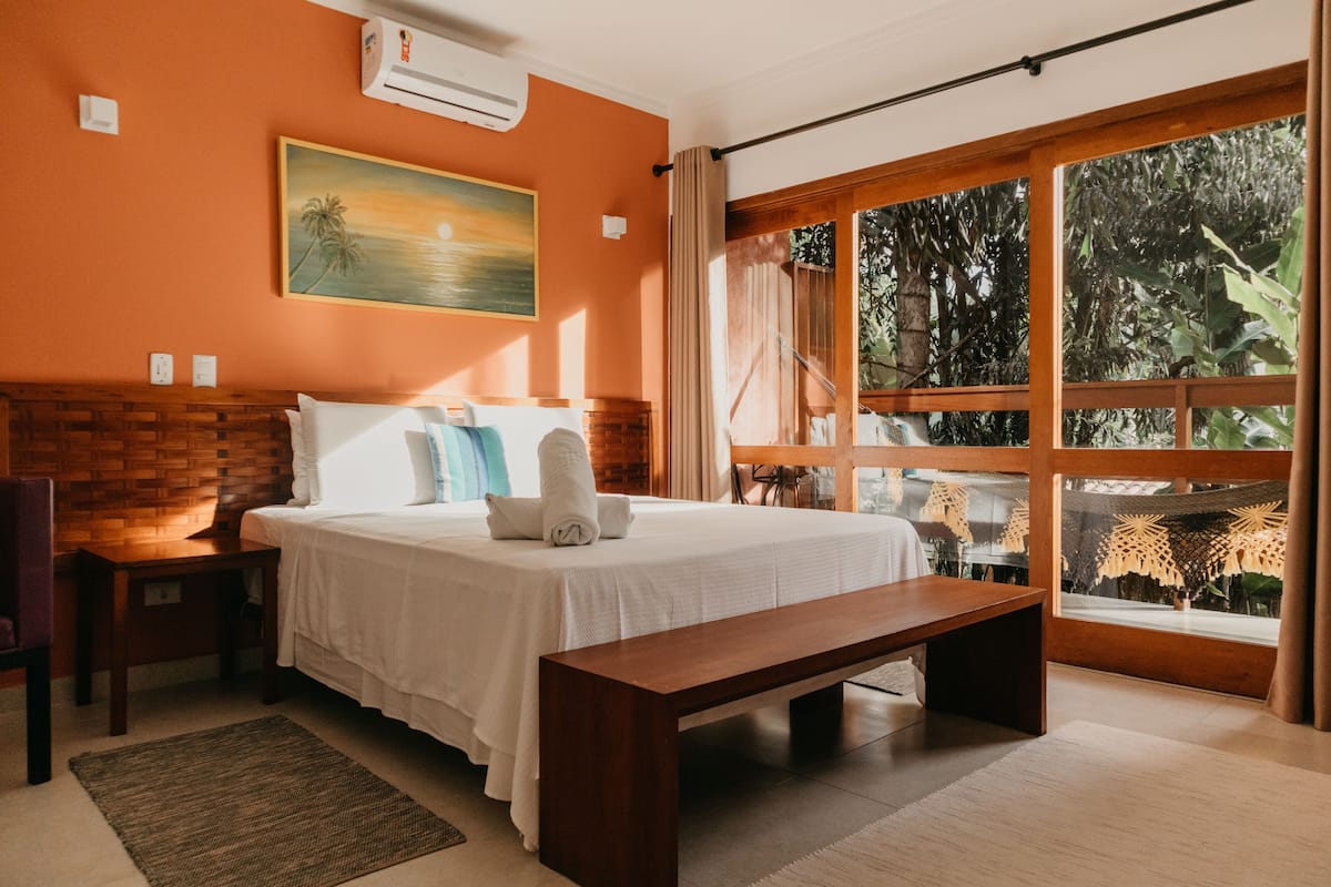 A bedroom with a wooden headboard, a large bed with white linens, a wooden bench, a window with a view of greenery, and an air conditioning unit on the wall.
