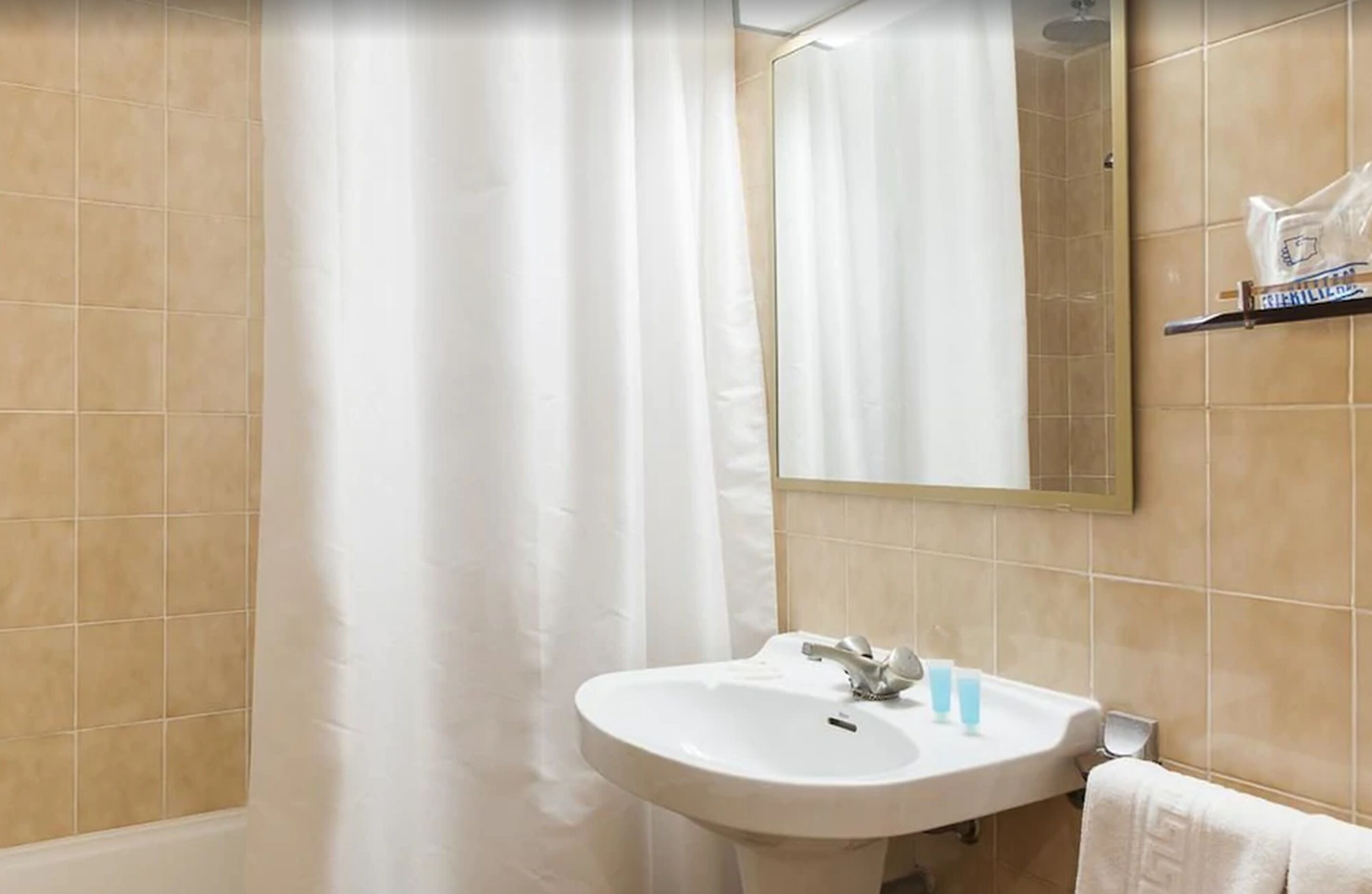 Une salle de bain avec des carreaux de couleur beige, un lavabo blanc, un miroir et un rideau de douche.