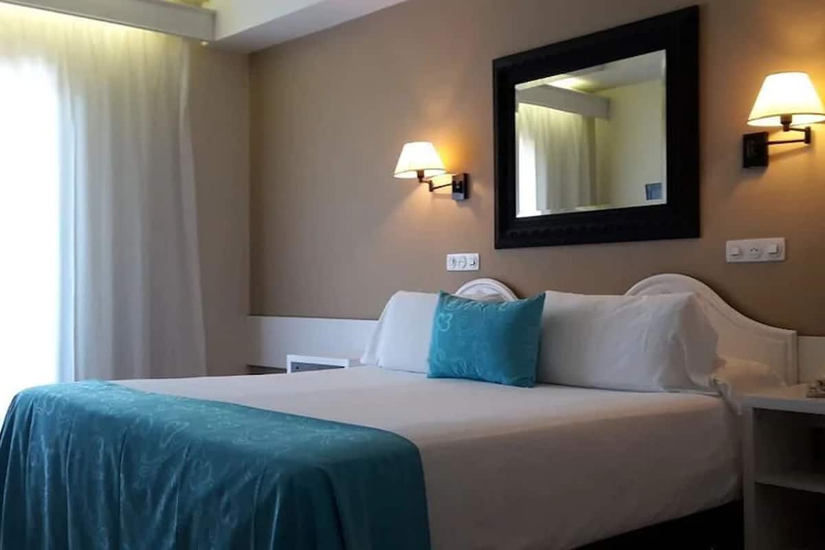 Quarto de hotel com cama, mesa de cabeceira, luminária, telefone e espelho na parede.