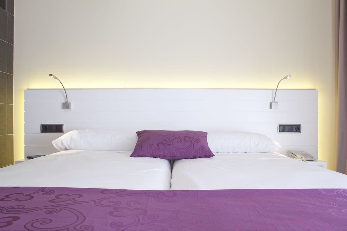 Uma cama de casal com lençóis brancos, um cobertor roxo e um travesseiro decorativo. Há duas mesas de cabeceira com abajures e um painel de controle fixado na parede.