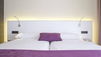 Een tweepersoonsbed met witte lakens, een paarse plaid en een decoratief kussen. Er zijn twee nachtkastjes met lampen en een bedieningspaneel aan de muur.