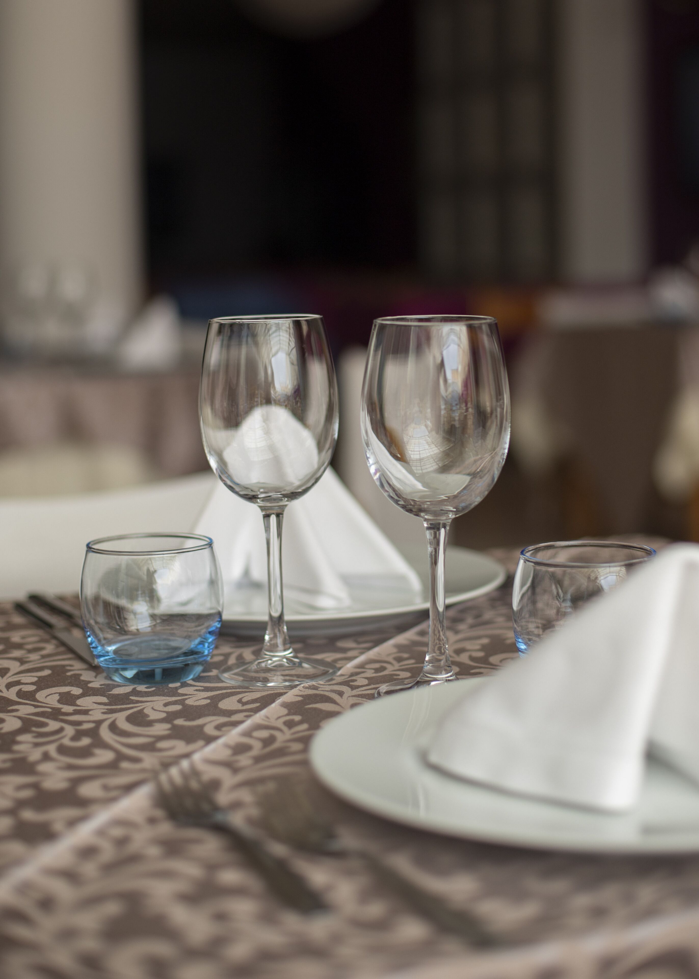 Une table à manger recouverte d’une nappe à motifs, avec une assiette blanche, une serviette blanche pliée et trois verres à vin.