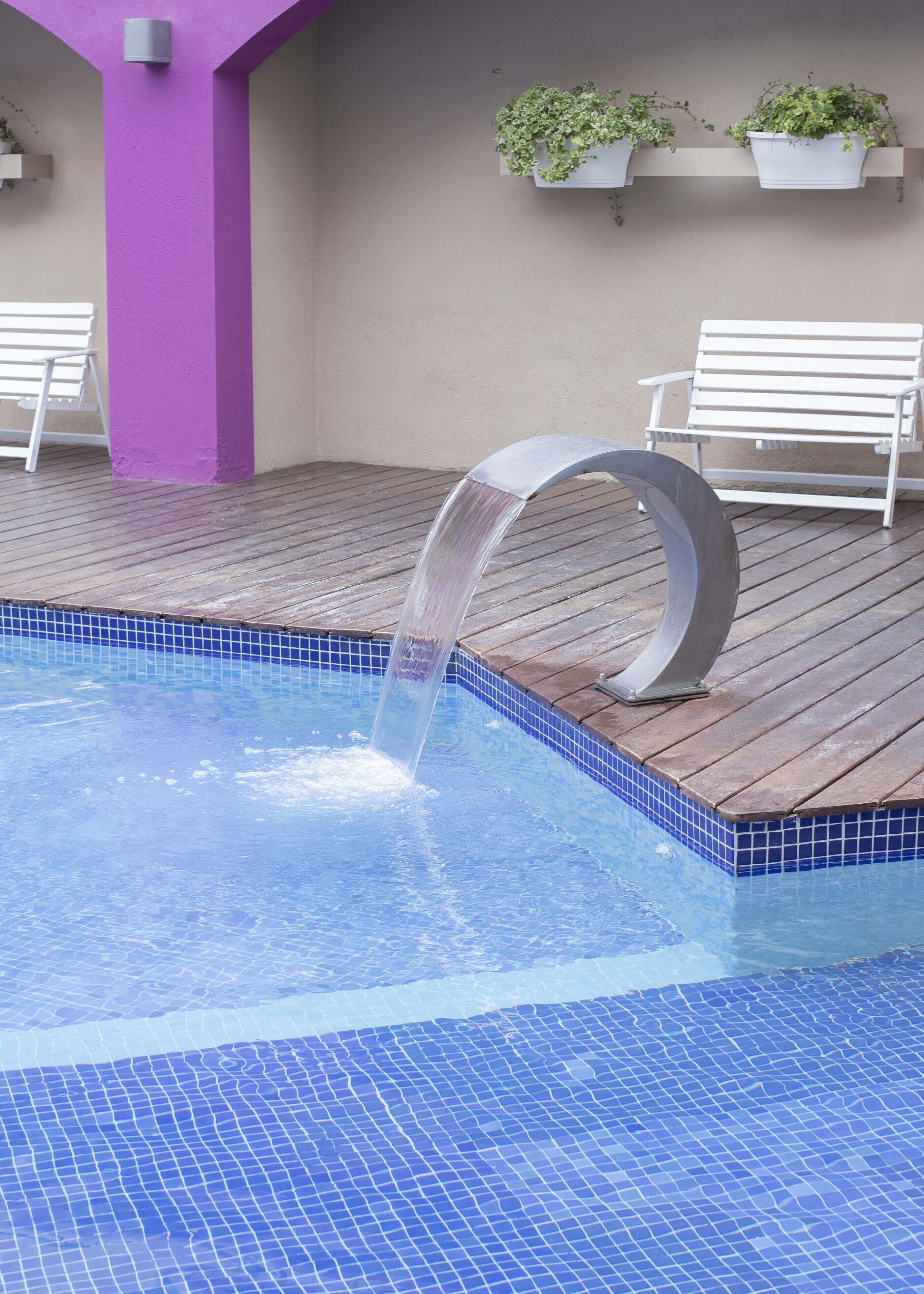 Une piscine agrémentée d’un élément décoratif aquatique, d’une terrasse en bois et de bancs blancs.