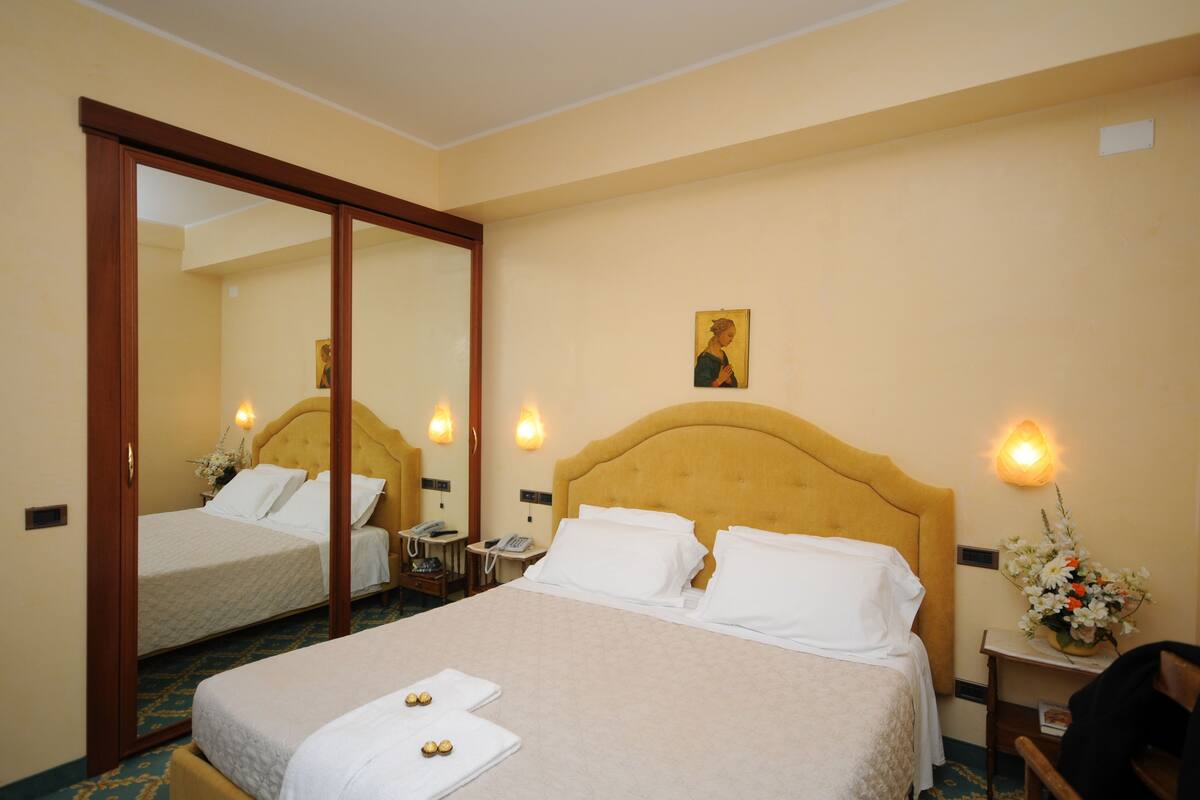 Une chambre d’hôtel avec deux lits, un grand miroir, des lampes de chevet, une petite table avec un téléphone et un arrangement floral décoratif.