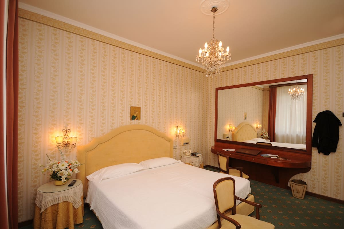 Une chambre d’hôtel avec un lit, un bureau, une chaise, un miroir et un lustre.