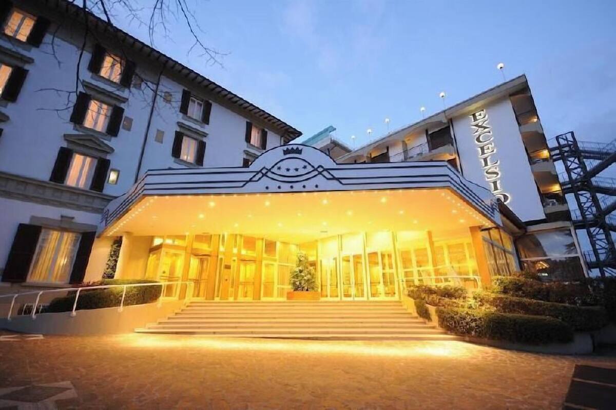 L’entrée d’un hôtel, avec un grand escalier, un emblème en forme de couronne et le nom « EXCELSIOR » inscrit sur la façade.