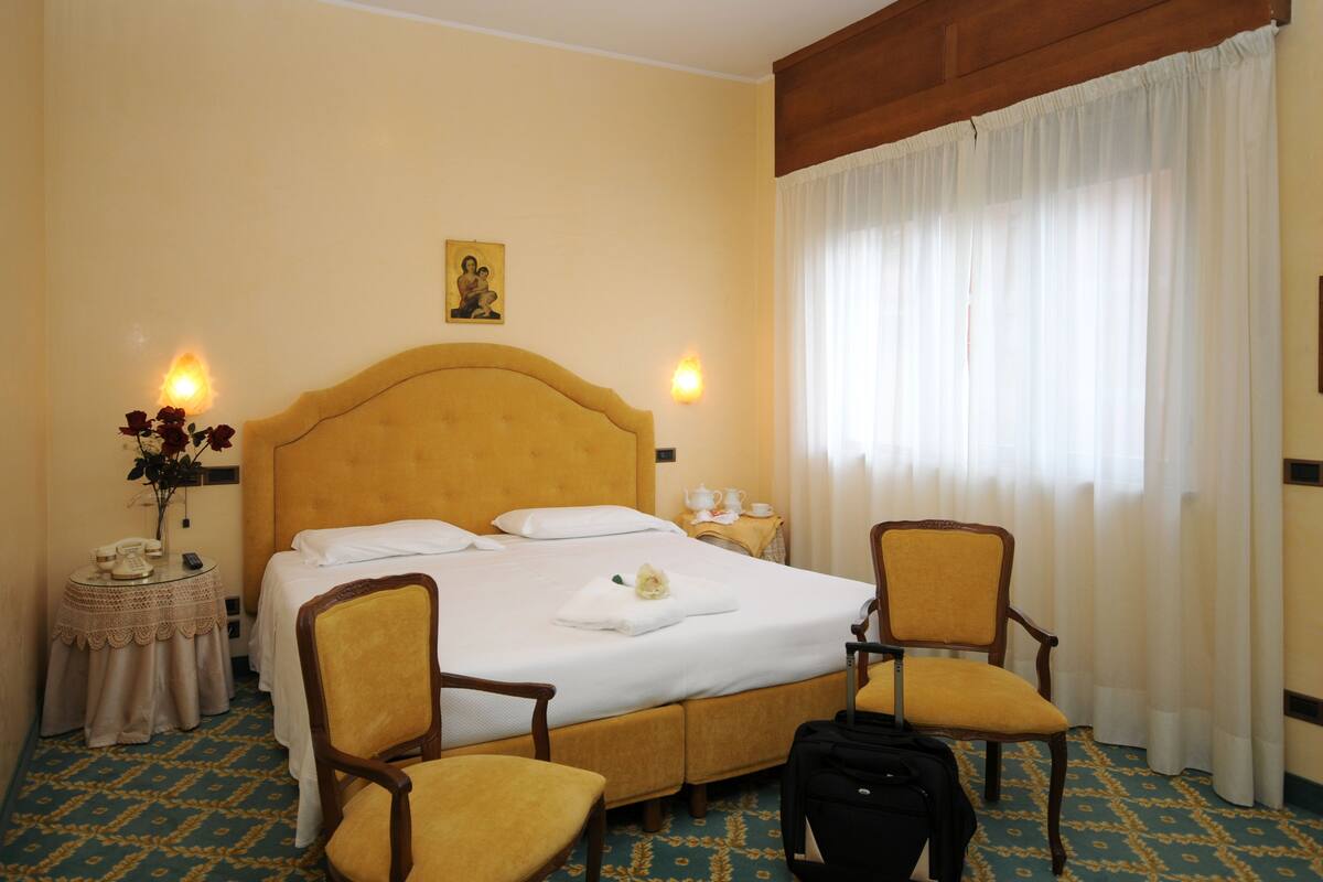 Une chambre d’hôtel avec un grand lit, deux chaises, une table de chevet avec une lampe et une fenêtre avec des rideaux.