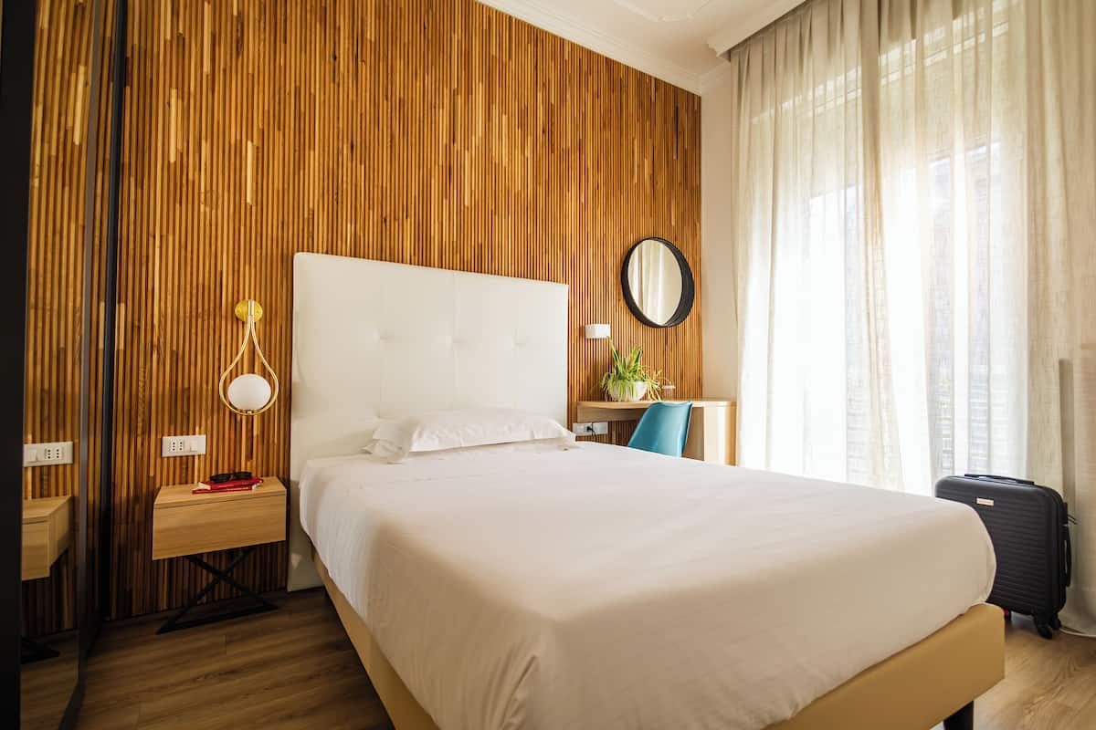 A hotel room with a large bed, a bedside table with a lamp, a desk with a chair, a mirror, and a window with curtains.