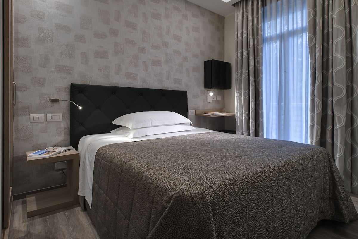 A hotel room with a large bed, a bedside table, a nightstand, a wall-mounted TV, and a window with curtains.