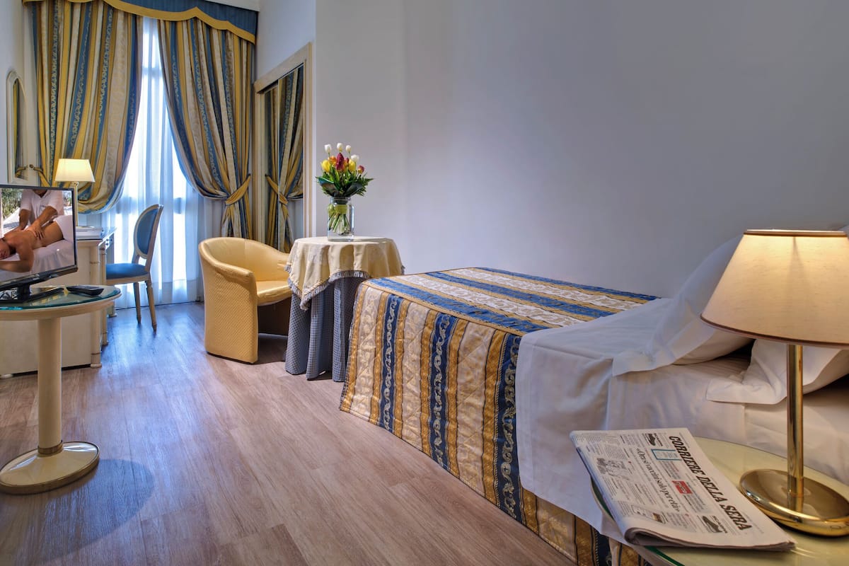 A hotel room with a bed, a desk with a computer, a chair, a table with a vase of flowers, and a lamp.