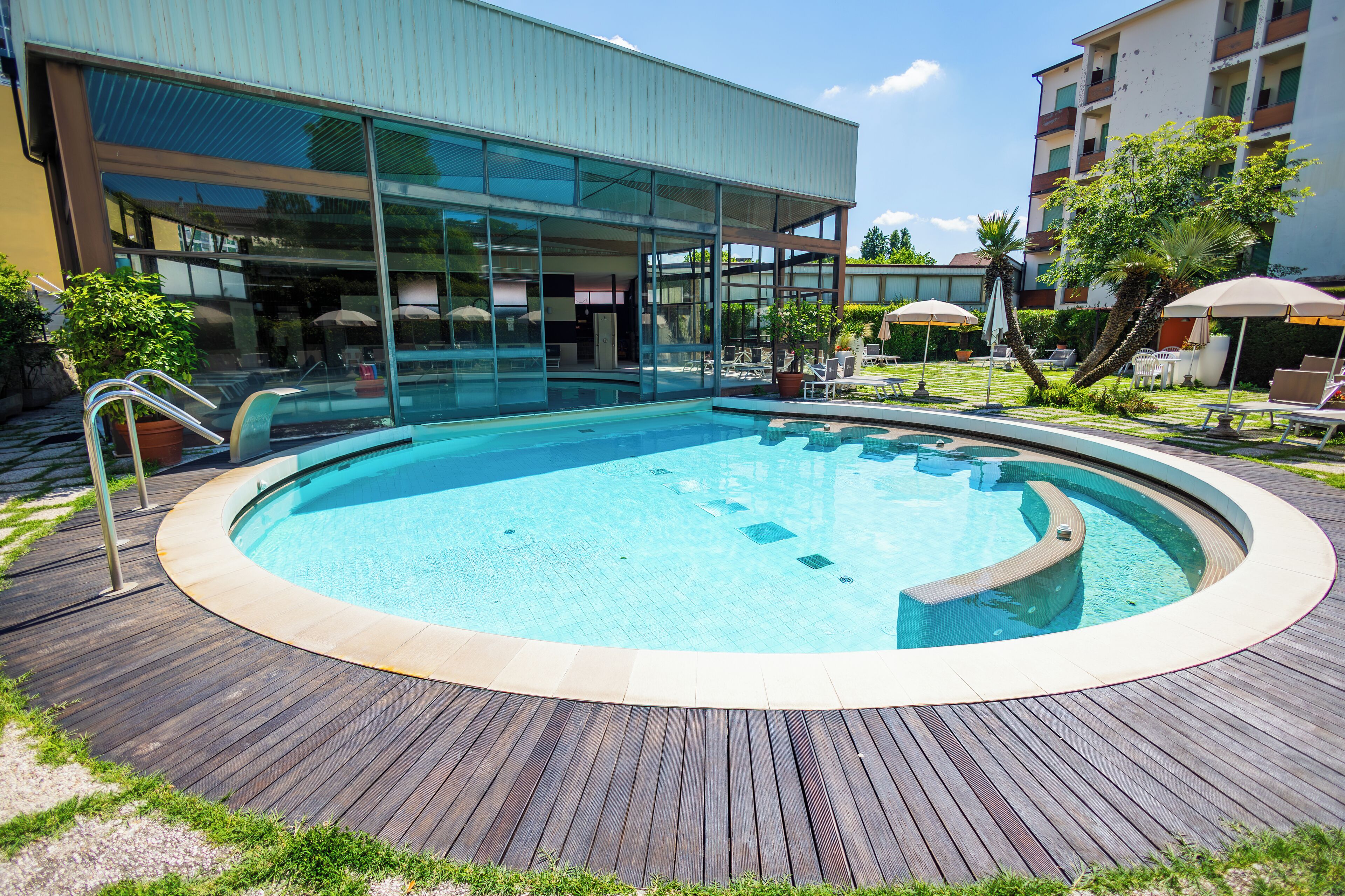 Un hotel moderno con piscina circondata da un solarium in legno, posti a sedere all'aperto e un edificio con pareti in vetro.