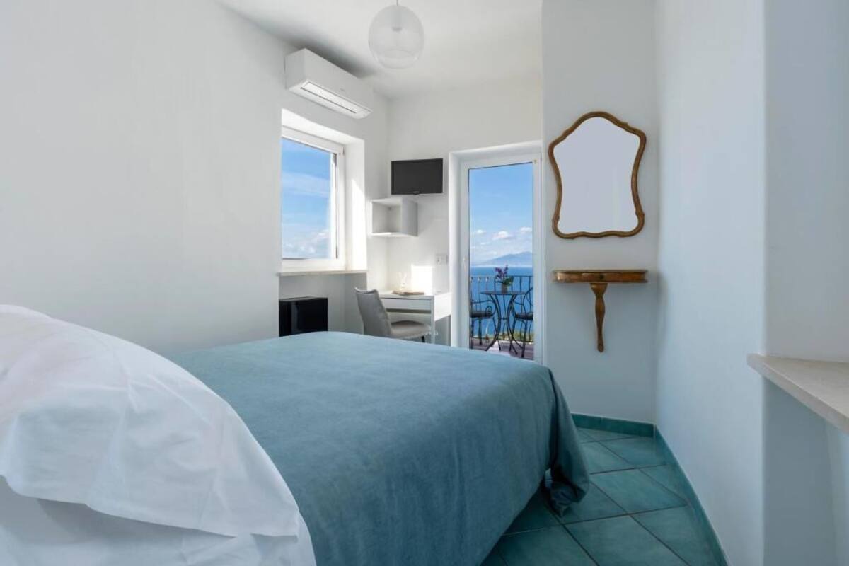 A bedroom with a bed, a desk, a chair, a TV, and a balcony with a view.