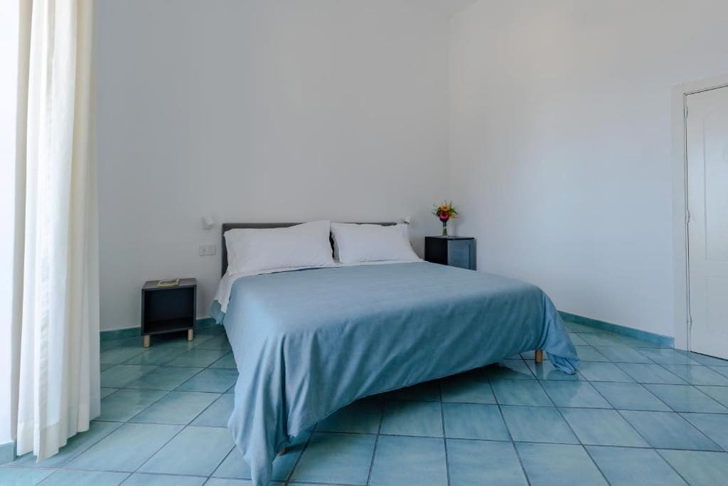 Quarto com cama, mesa de cabeceira, pequena mesa com um vaso e uma porta.