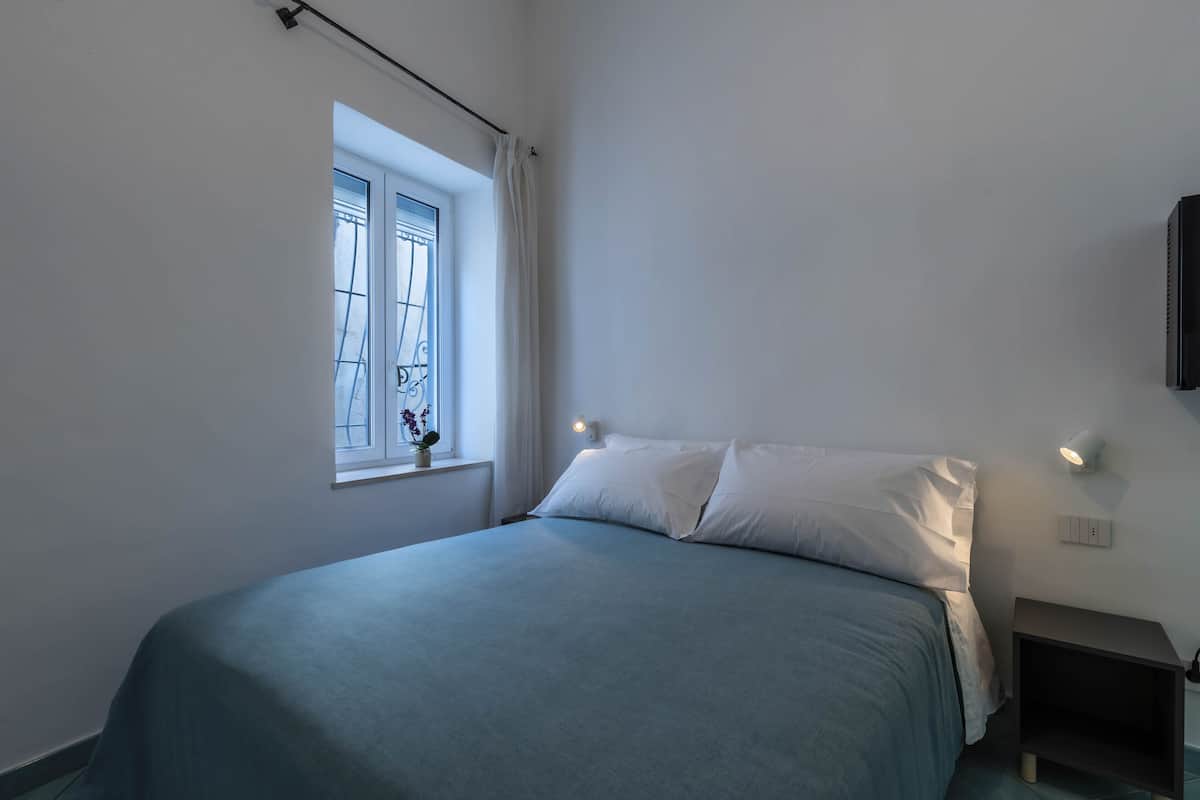 A bedroom with a bed, a window with curtains, a nightstand, and a wall-mounted light.