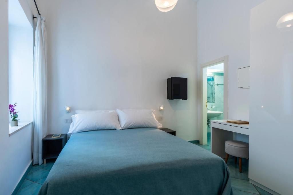 Een slaapkamer met een bed, nachtkastjes, een raam met gordijnen en een zichtbare badkamer door een open deur.