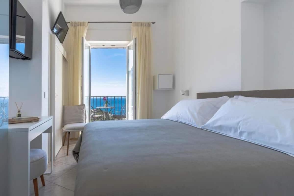 A hotel room with a bed, a desk, a chair, and a balcony with a view of the sea.