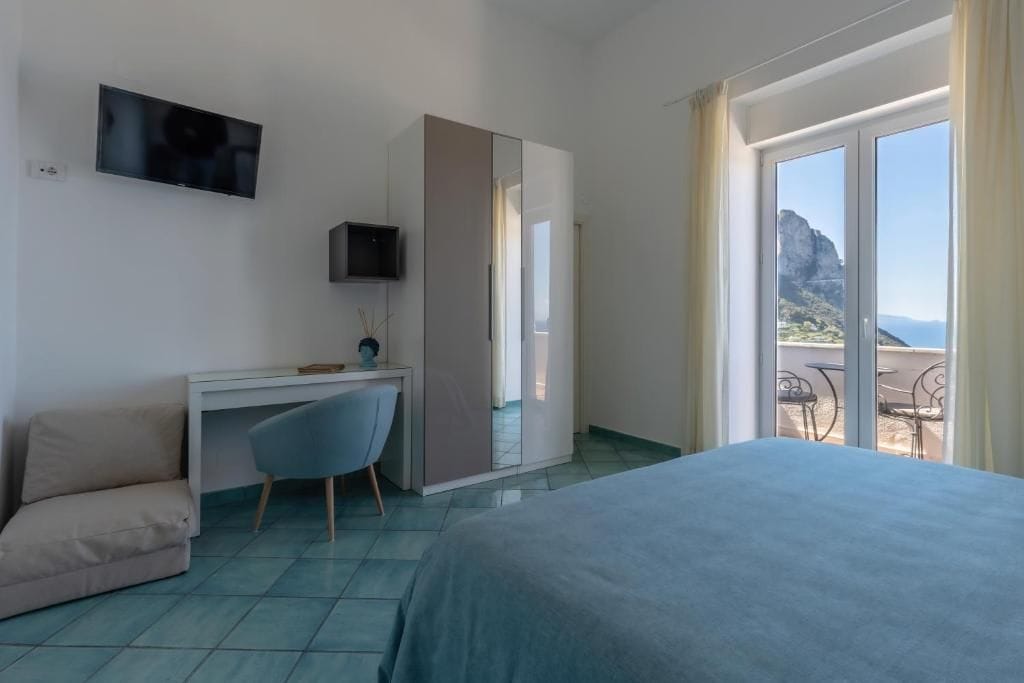 Habitación de hotel con cama, escritorio, silla, televisor y balcón con vistas.