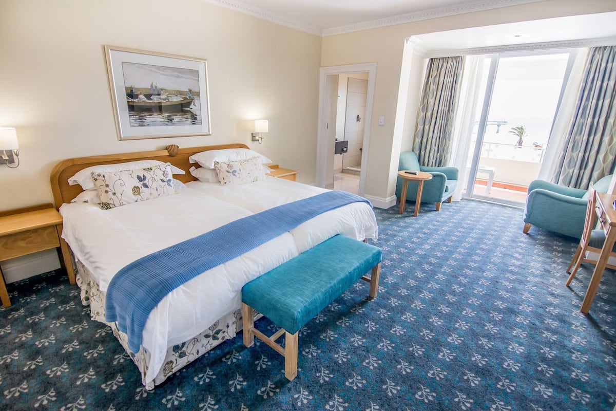 A hotel room with a large bed, a bench, a chair, a small table, a framed picture on the wall, and a view of the outside through a sliding door.