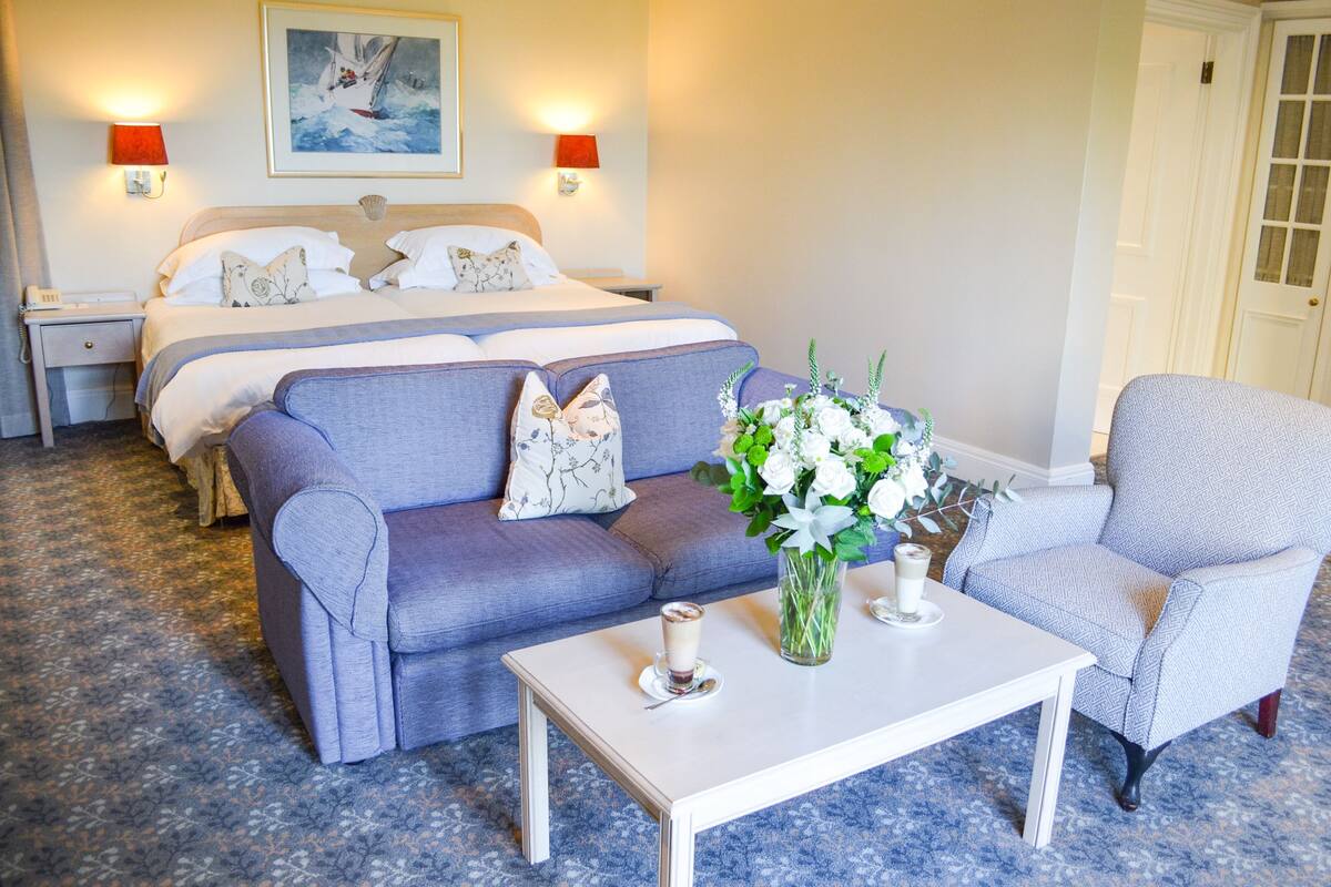 A hotel room with a bed, a sofa, a chair, a coffee table, and a vase with flowers.