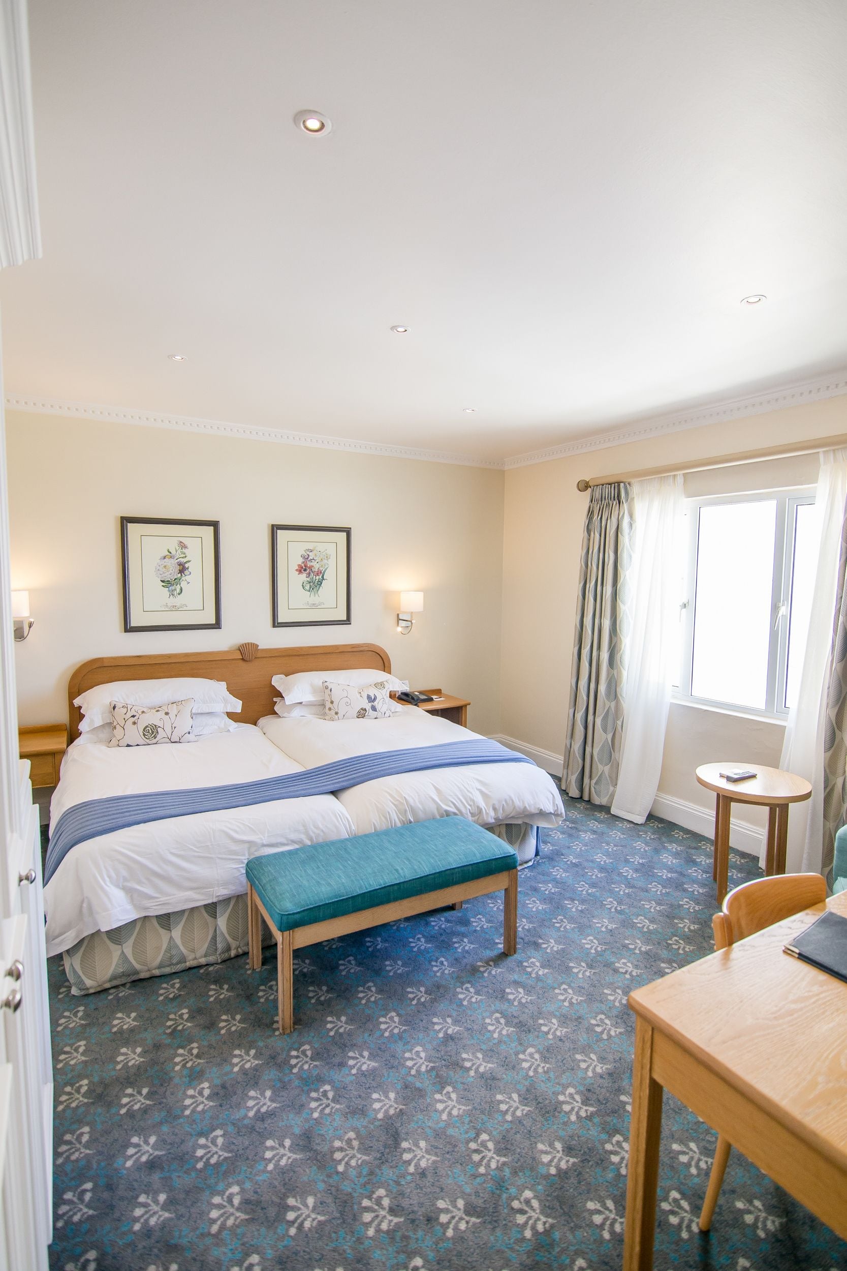 A hotel room with two beds, a bench, a small table, and a window with curtains.