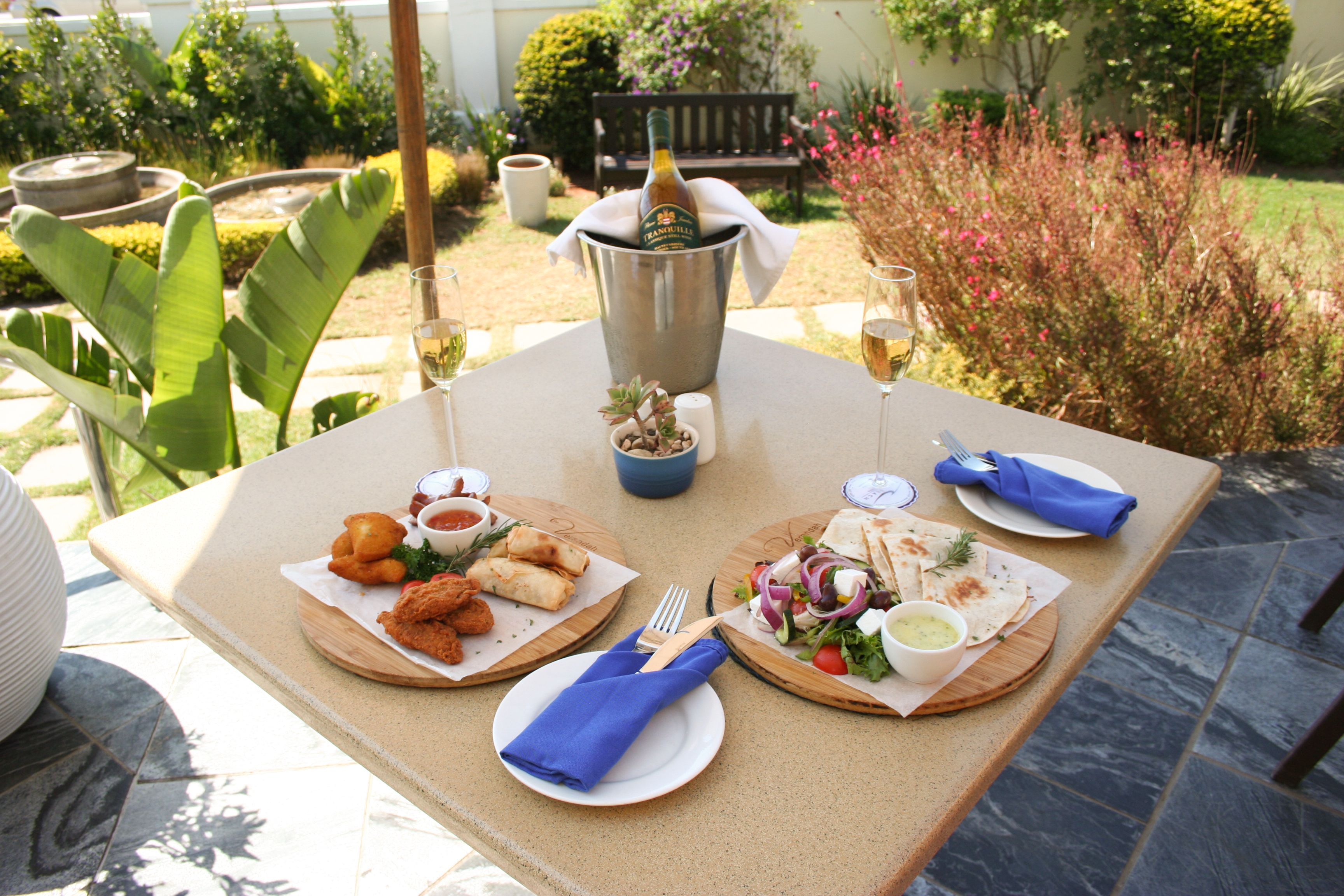 Una mesa preparada para una comida con varios platos, una botella de champán y vistas a un jardín.
