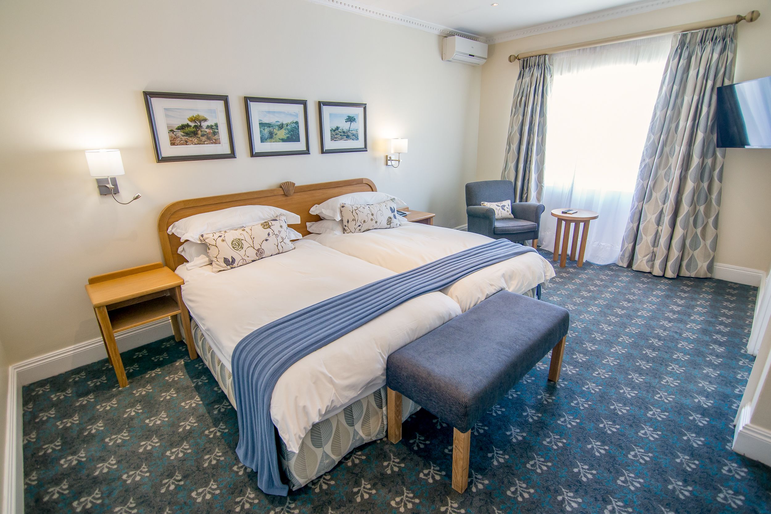 A hotel room with a large bed, a bedside table, a chair, a small table, and a TV mounted on the wall.