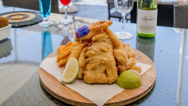 Un plateau de fruits de mer comprenant des produits frits et une tranche de citron vert, présenté sur une planche en bois, accompagné d’une bouteille de vin et d’une boisson bleue, le tout sur une table offrant une vue sur l’océan.