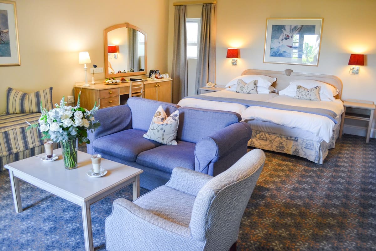 A hotel room with a bed, a sofa, a chair, a desk, and a table with a vase of flowers.