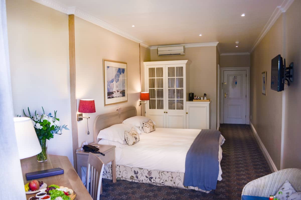A hotel room with a bed, bedside table, lamp, and a small desk with a telephone and a vase of flowers.