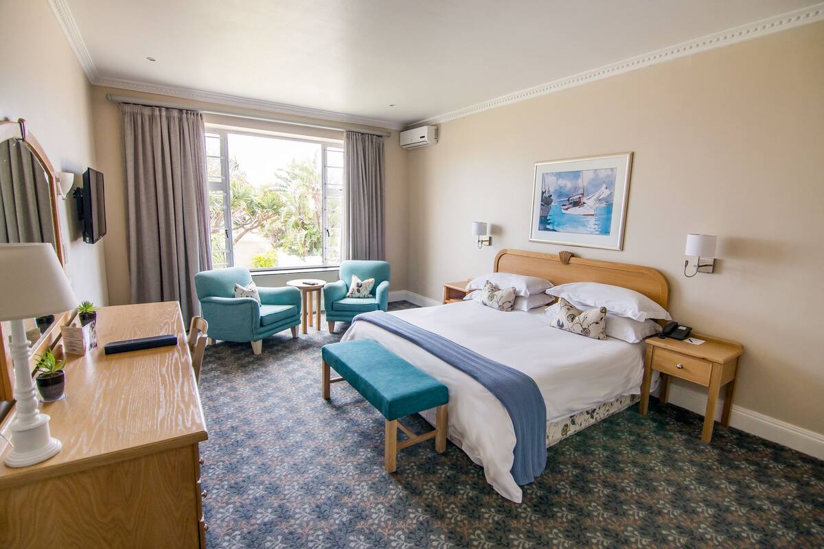 A hotel room with a bed, bedside tables, a desk, a chair, a bench, and a window with curtains.