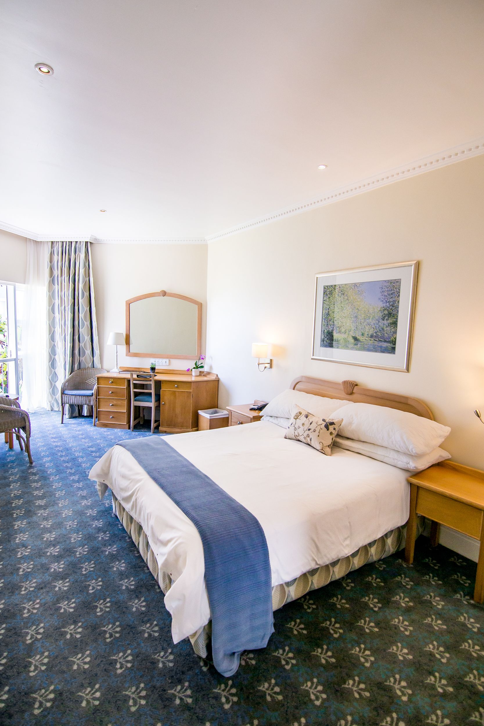 A hotel room with a large bed, a desk, a chair, a mirror, a nightstand, and a window with curtains.