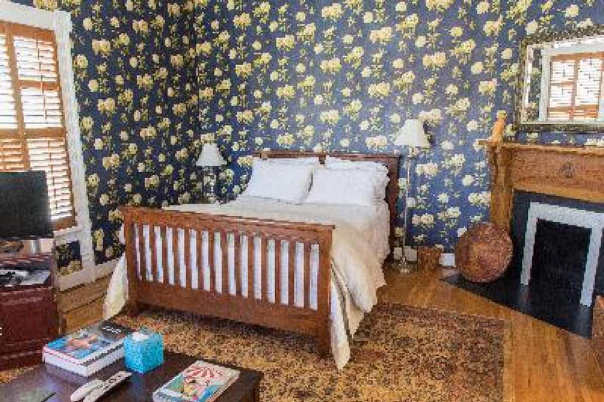 Une chambre à coucher avec un lit en bois, une table de chevet, une lampe, une cheminée et un papier peint à motifs floraux.