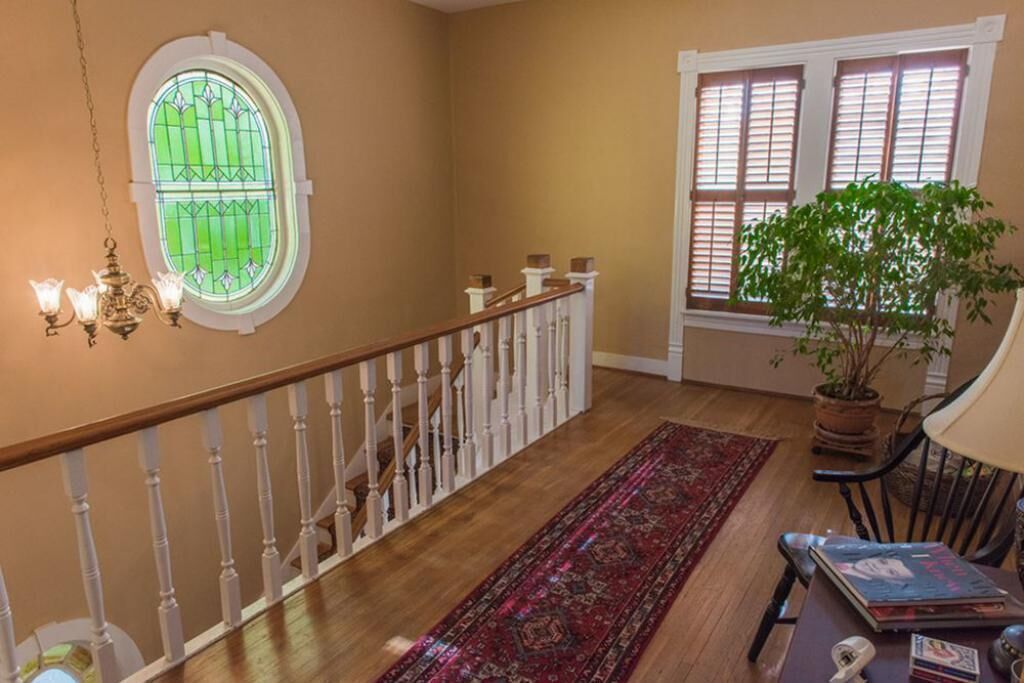 A staircase with a wooden handrail and white balusters, a red patterned rug, a potted plant, and a window with blinds.