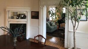 A dining area with a wooden table, chairs, and a vase with decorative branches. There is a fireplace with a mantel, a mirror, and a framed picture on the wall. A green sofa is visible in the background. - Charles Bass House Bed & Breakfast (South Boston)