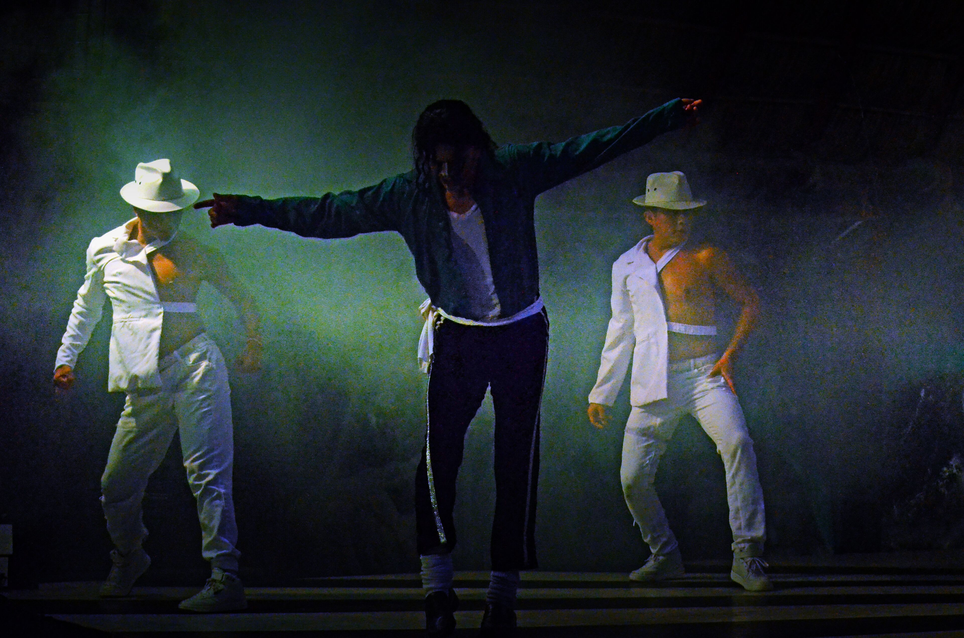 Un escenario con tres bailarines vestidos de blanco y sombreros, realizando una rutina de baile.