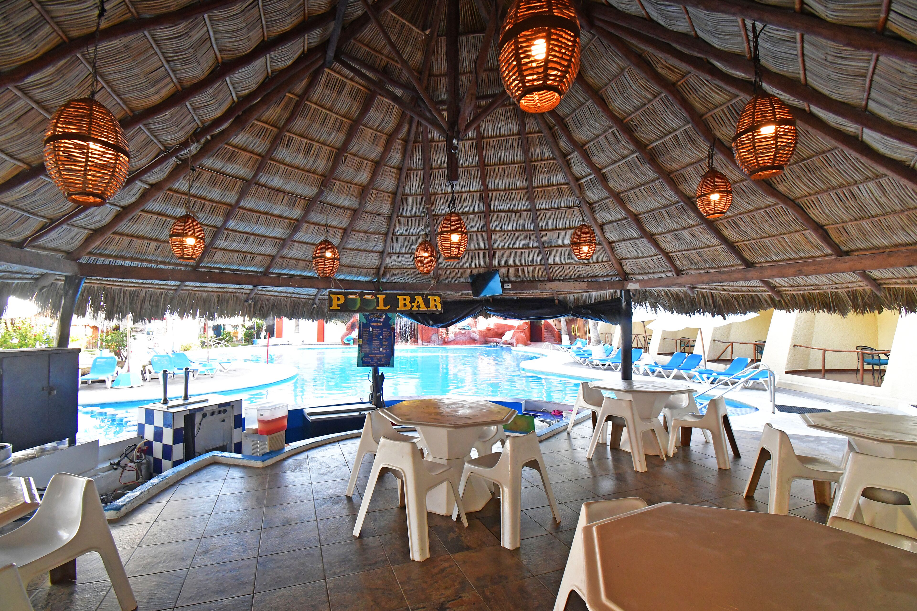 Zona de bar junto a la piscina con sillas y mesas de plástico blanco, techo de paja y lámparas de mimbre colgantes.
