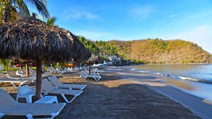 Une plage avec des chaises longues blanches et des parasols en chaume, un rivage sablonneux et, au loin, une colline couverte de végétation.