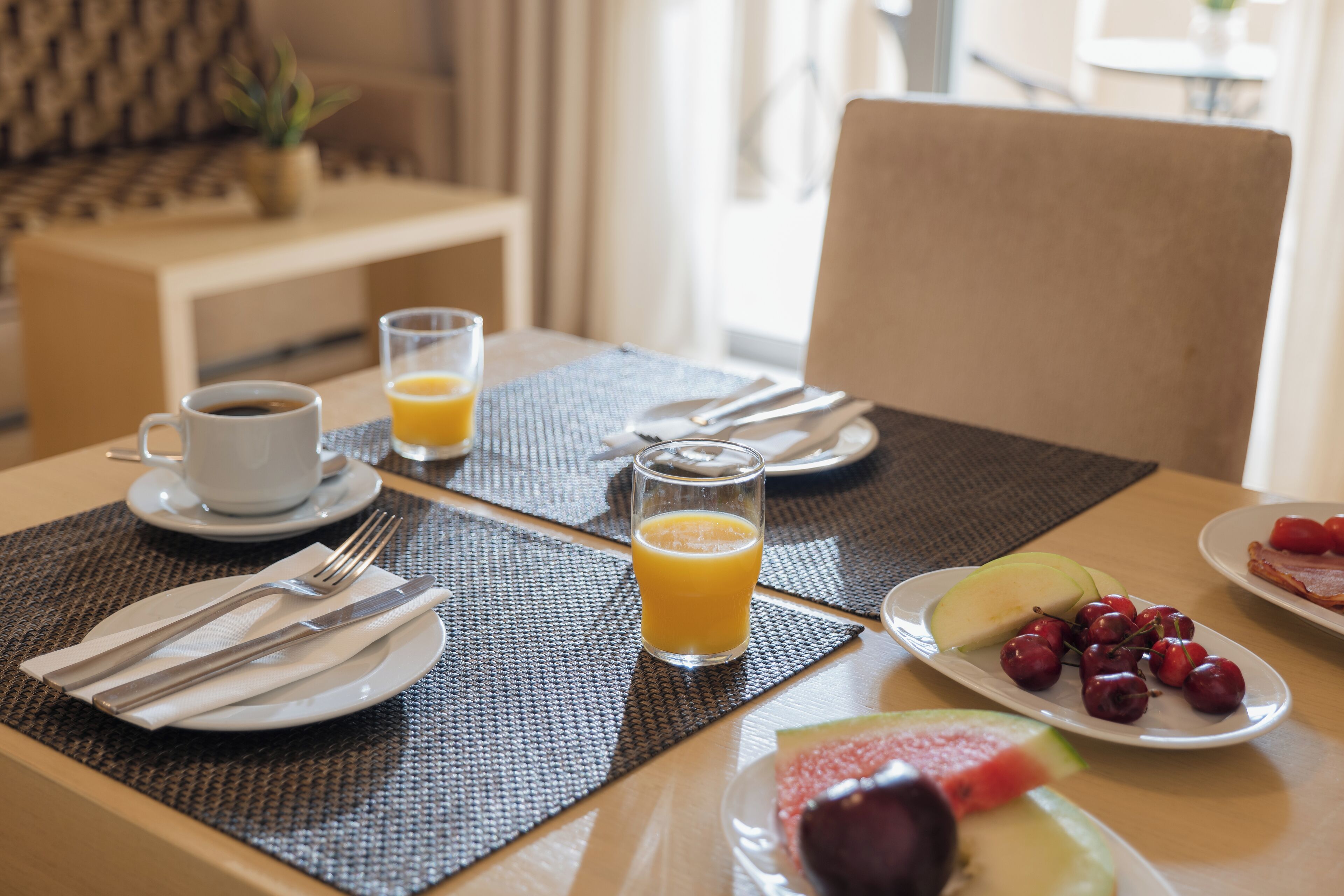 Une table mise pour le petit-déjeuner, avec une nappe pour deux personnes, comprenant des assiettes de fruits, un verre de jus d’orange et une tasse de café.
