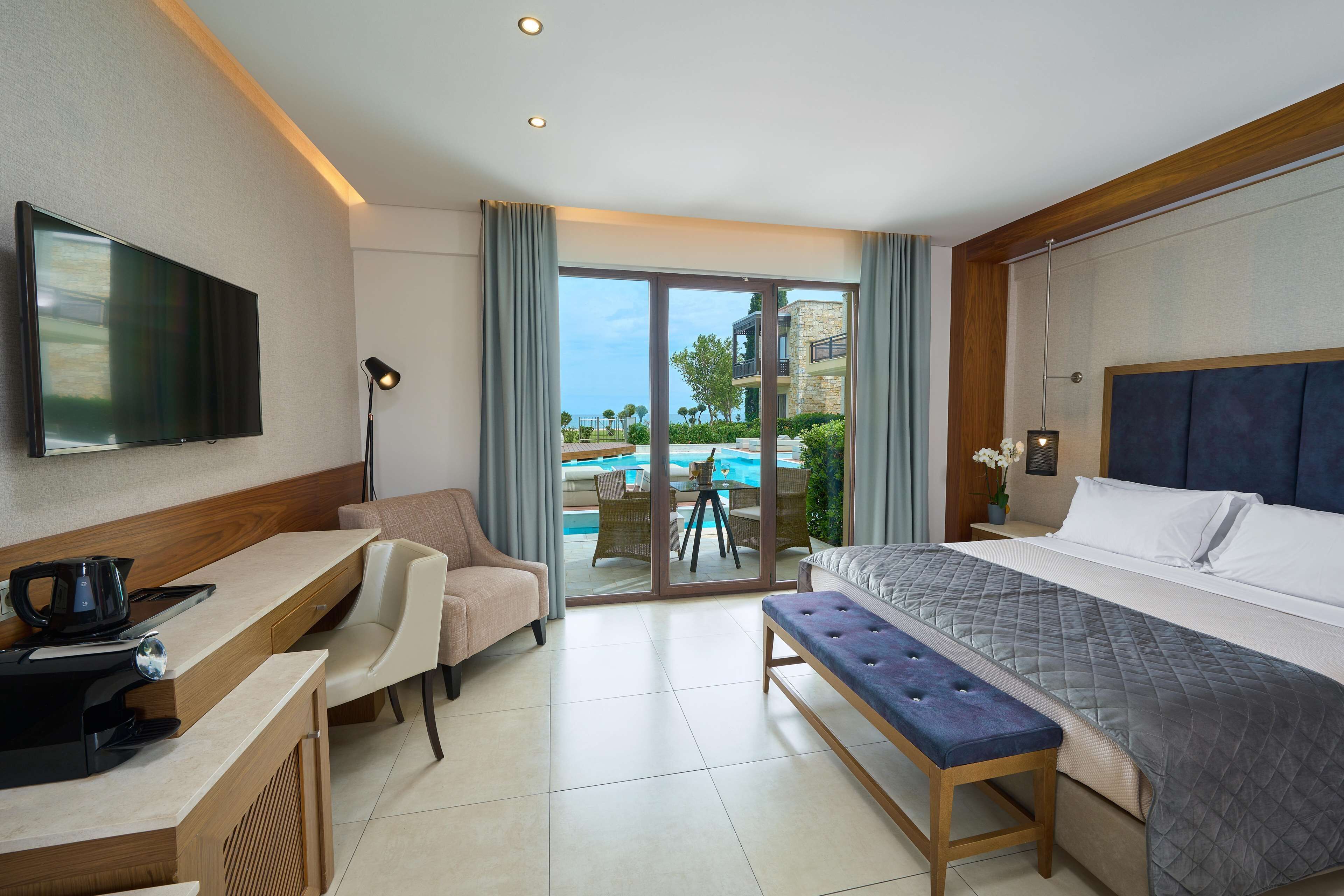 A hotel room with a bed, a desk with a computer, a chair, a bench, a TV, and a view of a pool and outdoor area.
